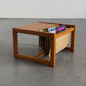 May include: A rectangular wooden side table with a smooth top surface and a built-in fabric sling magazine rack underneath. The frame is constructed from light brown wood, and the sling holds several magazines with visible IKEA branding.
