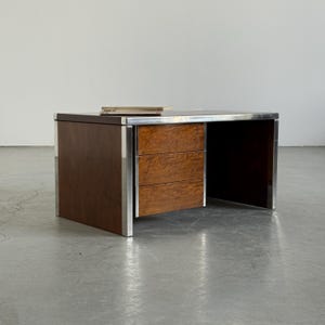 Op de afbeelding: Een rechthoekig bureau met een donkere houtnerfafwerking op de zijpanelen en een lichter, wortelhoutpatroon op de drie lades aan de voorzijde. De structuur is omlijst met een zilverkleurige metalen rand en heeft een vlak bovenblad.