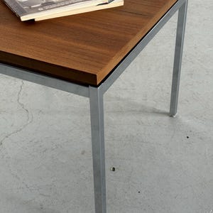 Op de afbeelding: Een salontafel in mid-century modern stijl met een rechthoekig, donkerbruin houten blad en een verchroomd metalen frame. Er ligt een tijdschrift op het tafelblad. De tafel is ongeveer 61 cm lang, 61 cm breed en 46 cm hoog.