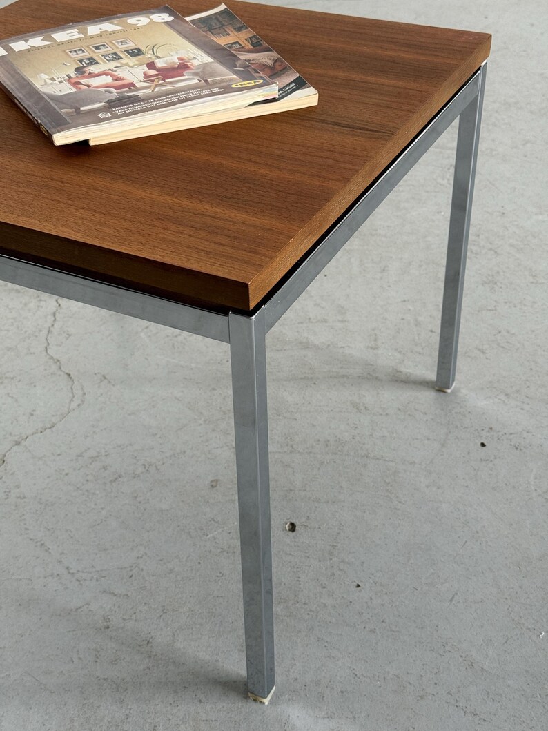 Op de afbeelding: Een salontafel in mid-century modern stijl met een rechthoekig, donkerbruin houten blad en een verchroomd metalen frame. Er ligt een tijdschrift op het tafelblad. De tafel is ongeveer 61 cm lang, 61 cm breed en 46 cm hoog.