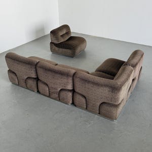 May include: A brown, mid-century modern sofa and armchair set. The sofa is a sectional with a geometric pattern, and the armchair has a rounded shape. Both pieces have a low profile and sit on metal legs.