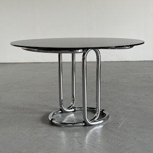 Puede incluir: Una mesa redonda con una superficie de cristal negro liso apoyada sobre una base de metal cromado pulido. La base consta de dos soportes tubulares verticales en forma de U conectados a un anillo circular en la parte inferior.