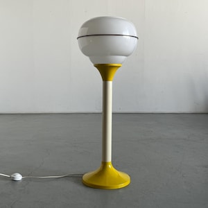 Puede incluir: Lámpara de mesa de estilo retro con una pantalla de globo blanca y una base amarilla. La lámpara tiene un tallo delgado de color blanquecino y un anillo negro alrededor de la pantalla. La base es de color amarillo brillante. La lámpara está enchufada.