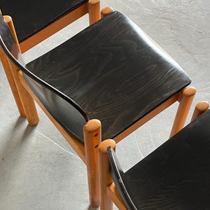 May include: Three wooden chairs with black seats and light brown legs. The chairs have a minimalist design, with rounded legs and a curved backrest. The seats are made of a dark wood with visible grain.