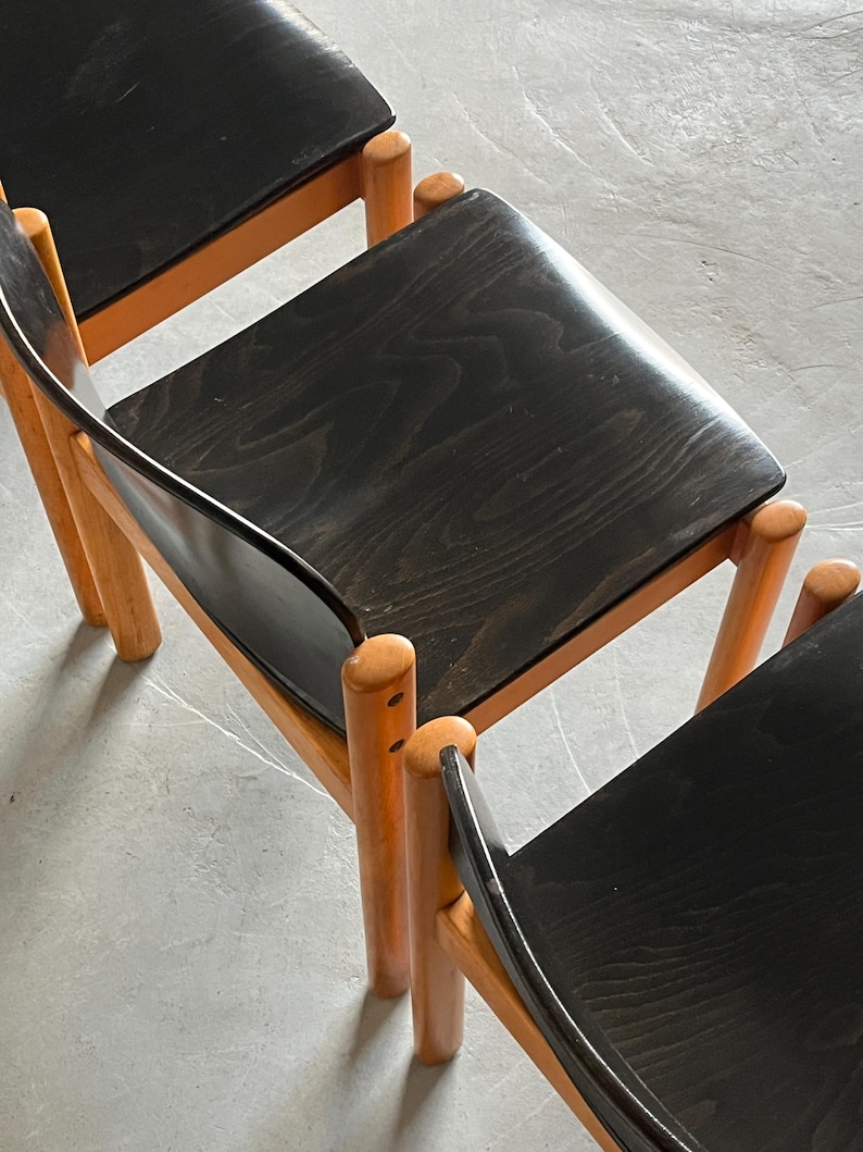 May include: Three wooden chairs with black seats and light brown legs. The chairs have a minimalist design, with rounded legs and a curved backrest. The seats are made of a dark wood with visible grain.