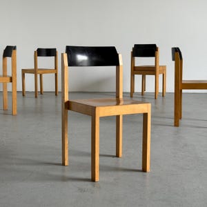 May include: A set of five wooden chairs with a minimalist design. The chairs feature light-colored wooden frames and square seats. The backs of the chairs are black and rectangular. The chairs are arranged in a room with a neutral background.