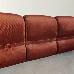 May include: Close-up view of a three-seater sofa's back. The sofa is upholstered in a rich, textured reddish-brown fabric.  Each section of the back is visibly padded and features a subtle seam detail. The fabric has a slightly ribbed or woven appearance. The sofa is positioned against a plain white wall, and the floor is a light gray concrete.