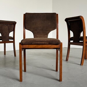 May include: Three wooden chairs with brown patterned fabric seats and backs. The chairs have a mid-century modern design, with straight legs and a curved backrest. The fabric has a small, repeating pattern.