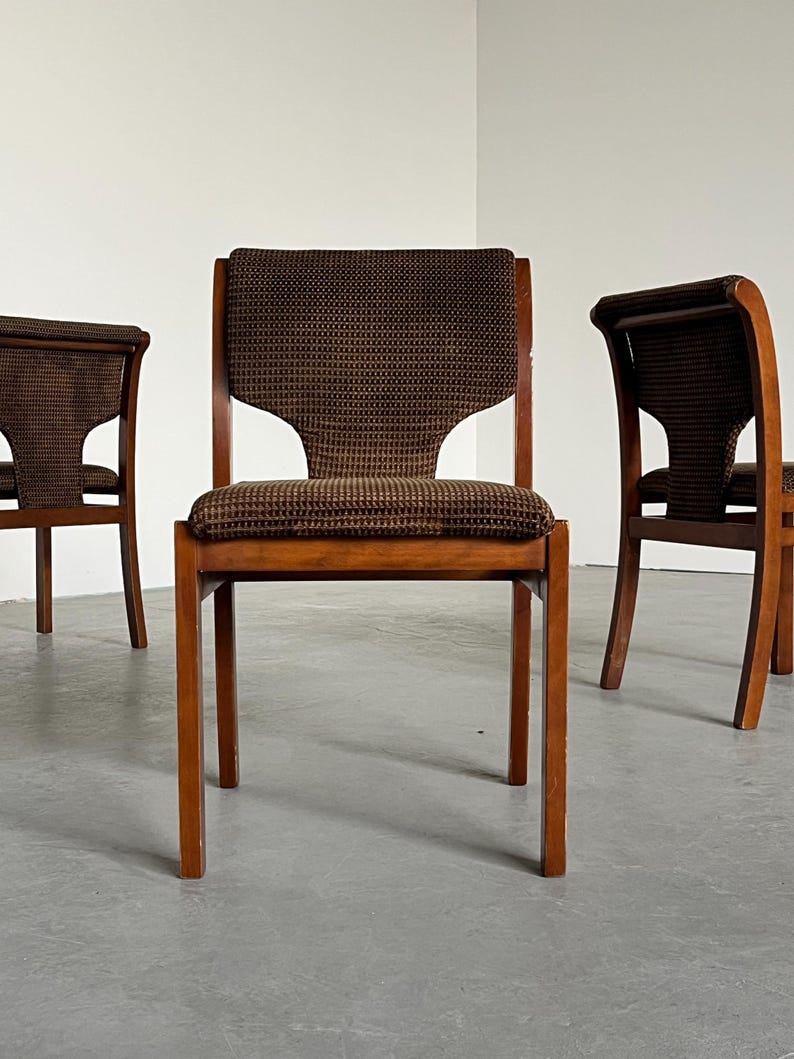 May include: Three wooden chairs with brown patterned fabric seats and backs. The chairs have a mid-century modern design, with straight legs and a curved backrest. The fabric has a small, repeating pattern.
