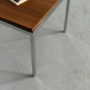 Op de afbeelding: Een salontafel in mid-century modern stijl met een rechthoekig, donkerhouten blad en chromen poten. Er ligt een tijdschrift op het tafelblad. De tafel is ongeveer 40 cm hoog.