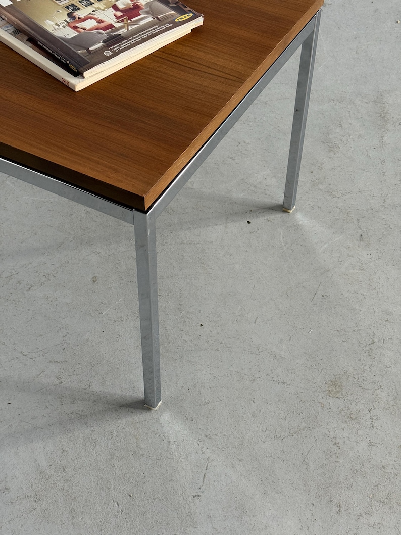 Op de afbeelding: Een salontafel in mid-century modern stijl met een rechthoekig, donkerhouten blad en chromen poten. Er ligt een tijdschrift op het tafelblad. De tafel is ongeveer 40 cm hoog.
