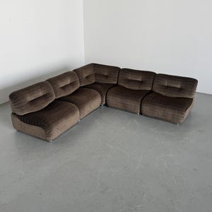May include: A brown sectional sofa with a curved design. The sofa is made of a textured fabric and has a low profile. The sofa is composed of multiple modular pieces, creating an L-shape. The sofa sits on a silver metal base.