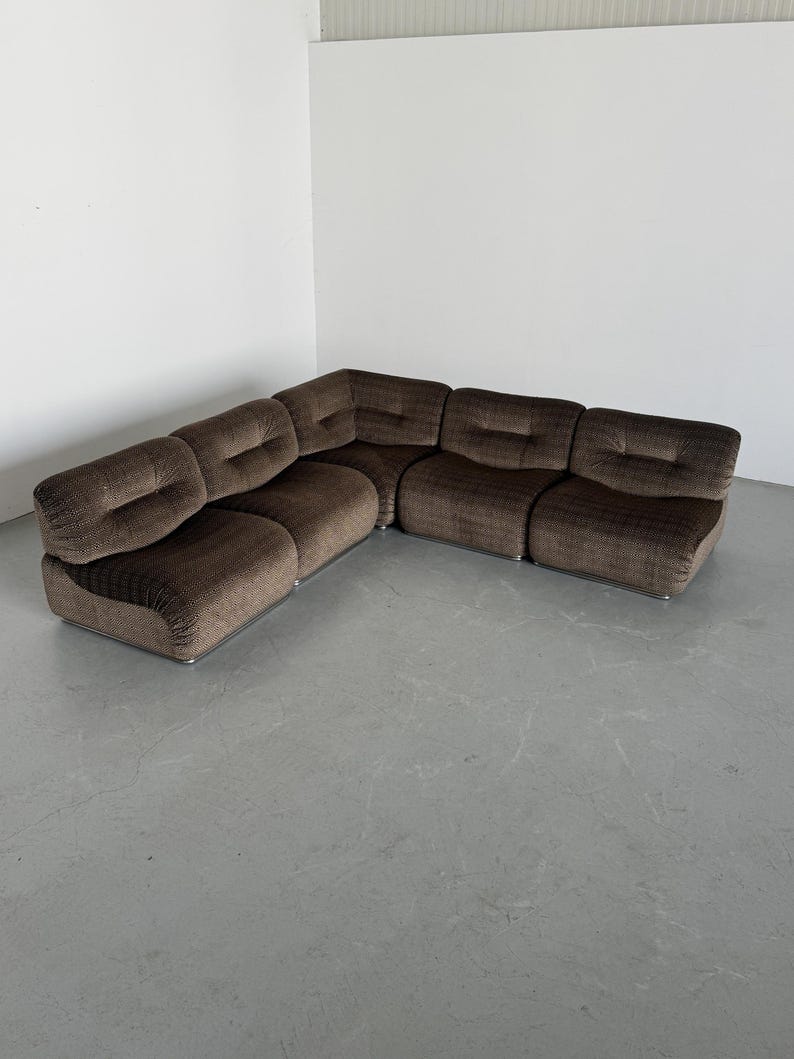 May include: A brown sectional sofa with a curved design. The sofa is made of a textured fabric and has a low profile. The sofa is composed of multiple modular pieces, creating an L-shape. The sofa sits on a silver metal base.