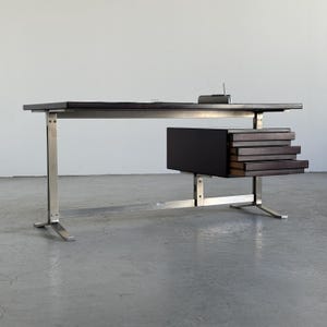 May include: A rectangular desk featuring a dark brown wooden top and a matching three-drawer unit suspended on the right side. The frame is constructed from brushed silver metal with H-shaped legs and a horizontal support bar connecting the base.