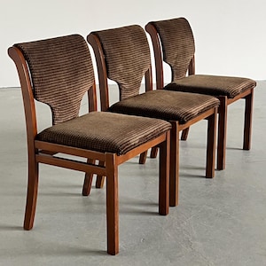 May include: Three wooden chairs with brown patterned fabric seats and backs. The chairs have a classic design with curved backs and straight legs. The wood has a warm, reddish-brown tone, and the fabric has a textured appearance.
