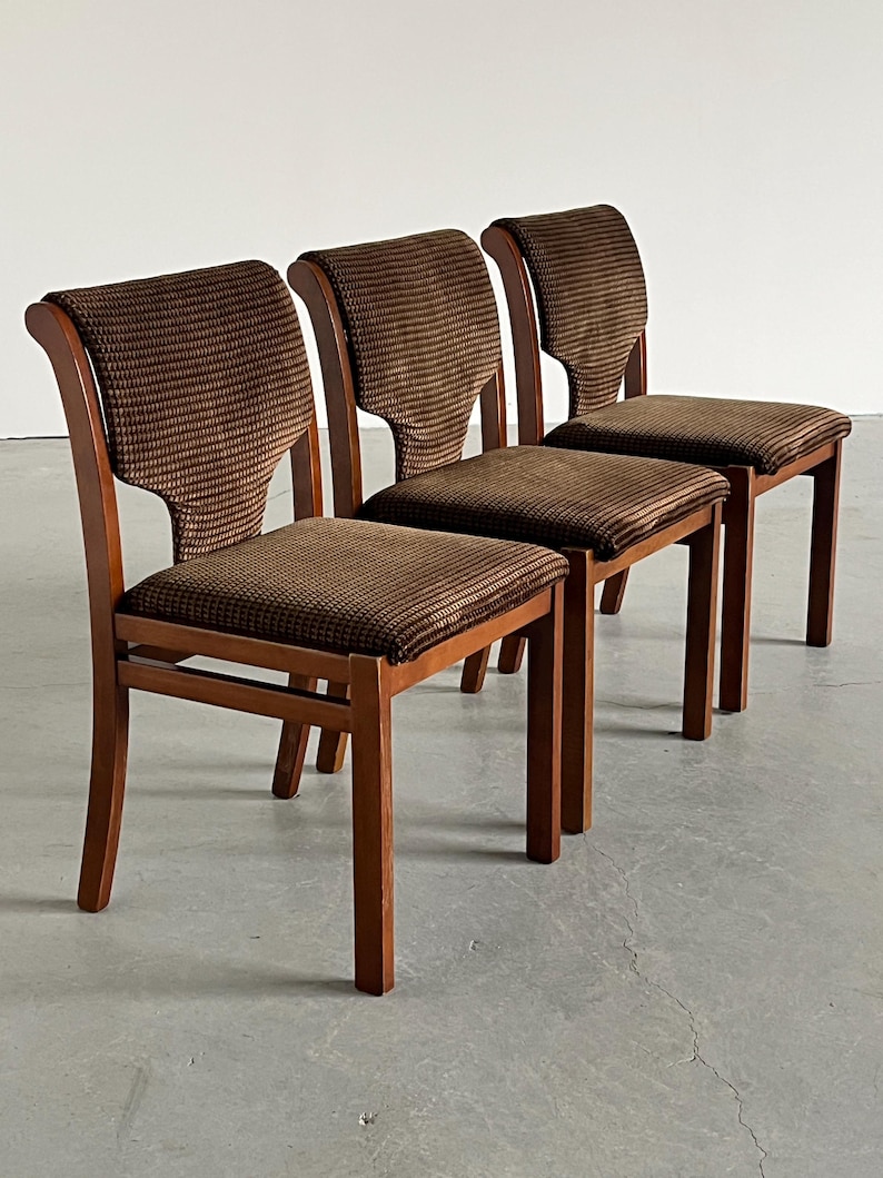 May include: Three wooden chairs with brown patterned fabric seats and backs. The chairs have a classic design with curved backs and straight legs. The wood has a warm, reddish-brown tone, and the fabric has a textured appearance.