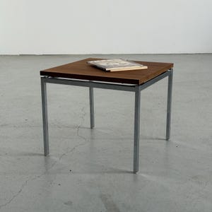 Op de afbeelding: Een vierkante bijzettafel met een donkerbruin houten blad en een zilverkleurig metalen frame. Er ligt een boek op de tafel. De tafel is ongeveer 46 cm vierkant en 46 cm hoog.