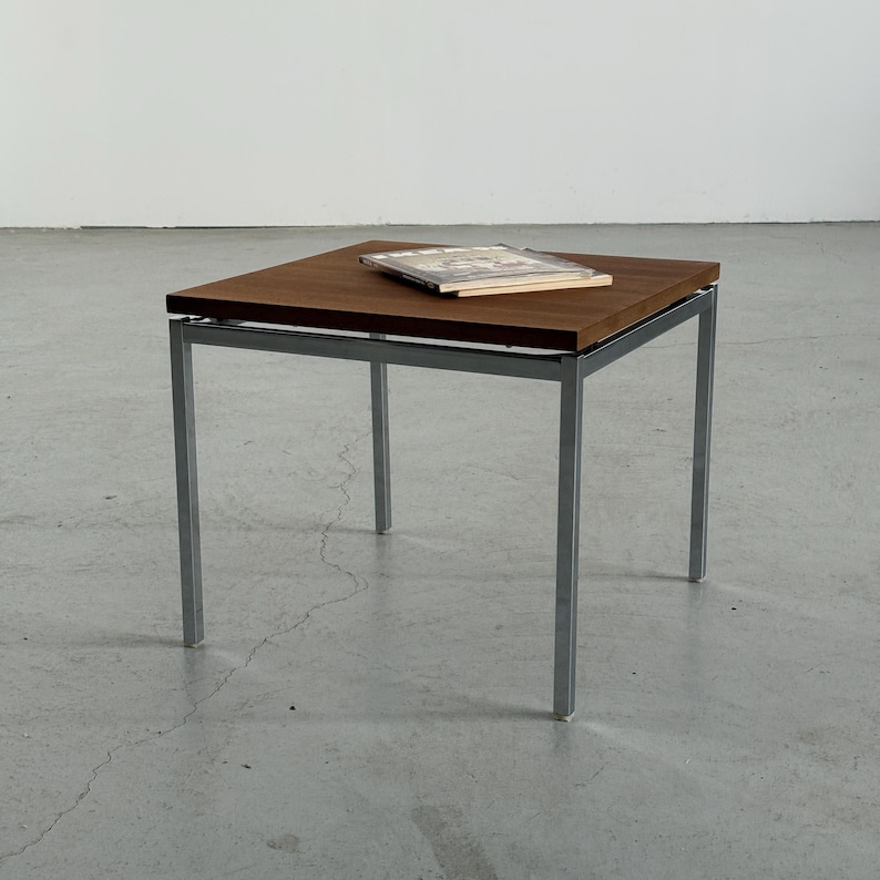 Op de afbeelding: Een vierkante bijzettafel met een donkerbruin houten blad en een zilverkleurig metalen frame. Er ligt een boek op de tafel. De tafel is ongeveer 46 cm vierkant en 46 cm hoog.