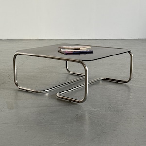 May include: A square coffee table featuring a dark, reflective glass top supported by a continuous, tubular chrome metal frame. The frame forms a distinct, rounded geometric base that curves upward to hold the tabletop. A small stack of magazines rests on top.