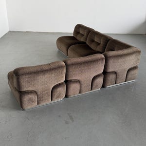 May include: Brown modular sofa with a geometric pattern. The sofa has a low profile with a rounded design and a silver metal base. The cushions are tufted, and the sofa is arranged in an L-shape. The sofa is set against a plain white wall.