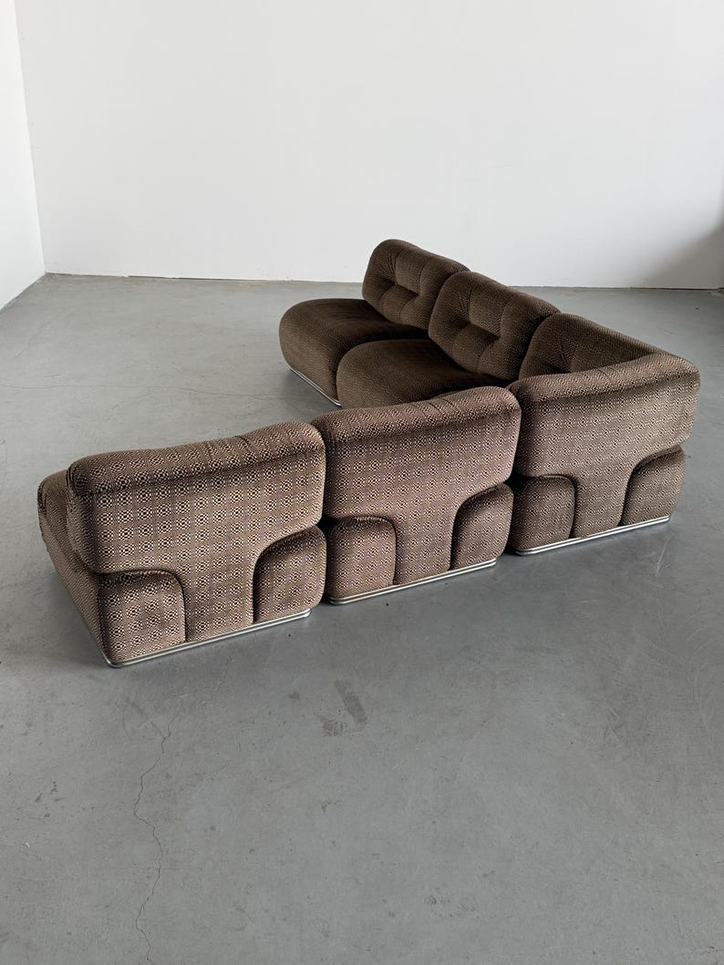 May include: Brown modular sofa with a geometric pattern. The sofa has a low profile with a rounded design and a silver metal base. The cushions are tufted, and the sofa is arranged in an L-shape. The sofa is set against a plain white wall.