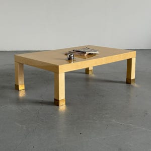 Peut inclure: Table basse rectangulaire avec une finition beige clair et des accents en laiton sur les pieds. La table a une surface brillante et contient un petit verre et une pile de magazines. La table mesure environ 122 cm de long.