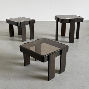 Puede incluir: Tres mesas auxiliares cuadradas de color marrón oscuro con tapas de cristal. Las mesas tienen un diseño minimalista con patas rectangulares gruesas y un acabado de madera oscura. Las tapas de cristal están tintadas y las mesas están dispuestas sobre un suelo de color neutro.