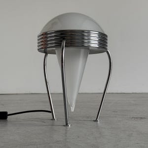 May include: A table lamp featuring a white frosted glass dome top and a matching conical glass base. The structure is supported by three curved chrome metal legs and includes a ribbed chrome band encircling the center of the lamp body.