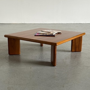Puede incluir: Una mesa de centro cuadrada de madera con una superficie marrón lisa y pulida con bordes redondeados. La mesa cuenta con cuatro patas gruesas de panel plano situadas en cada esquina, que sostienen la parte superior baja. Dos revistas descansan en el centro.