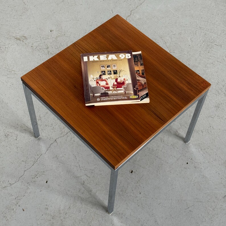 Op de afbeelding: Een vierkante salontafel met een donkerhouten blad en chromen poten. Een "IKEA 98" catalogus ligt op de tafel, met interieurontwerp. De tafel is ongeveer 61 cm vierkant.