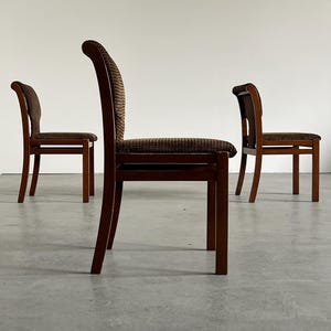 May include: Three wooden dining chairs with dark brown frames and patterned seat cushions. The chairs have curved backs and are arranged in a room with a neutral background. The chairs are made of wood.
