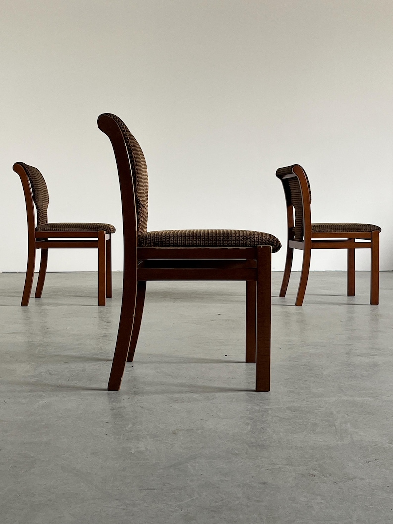 May include: Three wooden dining chairs with dark brown frames and patterned seat cushions. The chairs have curved backs and are arranged in a room with a neutral background. The chairs are made of wood.