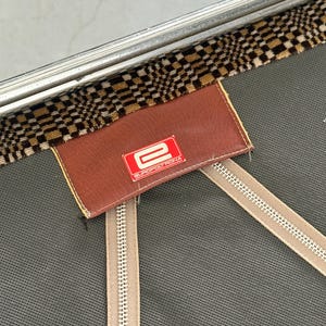 May include: Close-up of a vintage suitcase detail. The image shows a brown leather tag with the red "E" logo of EUROPOLTRONA. The tag is stitched onto a gray fabric with two beige zippers. A checkered brown and tan fabric trim is visible.