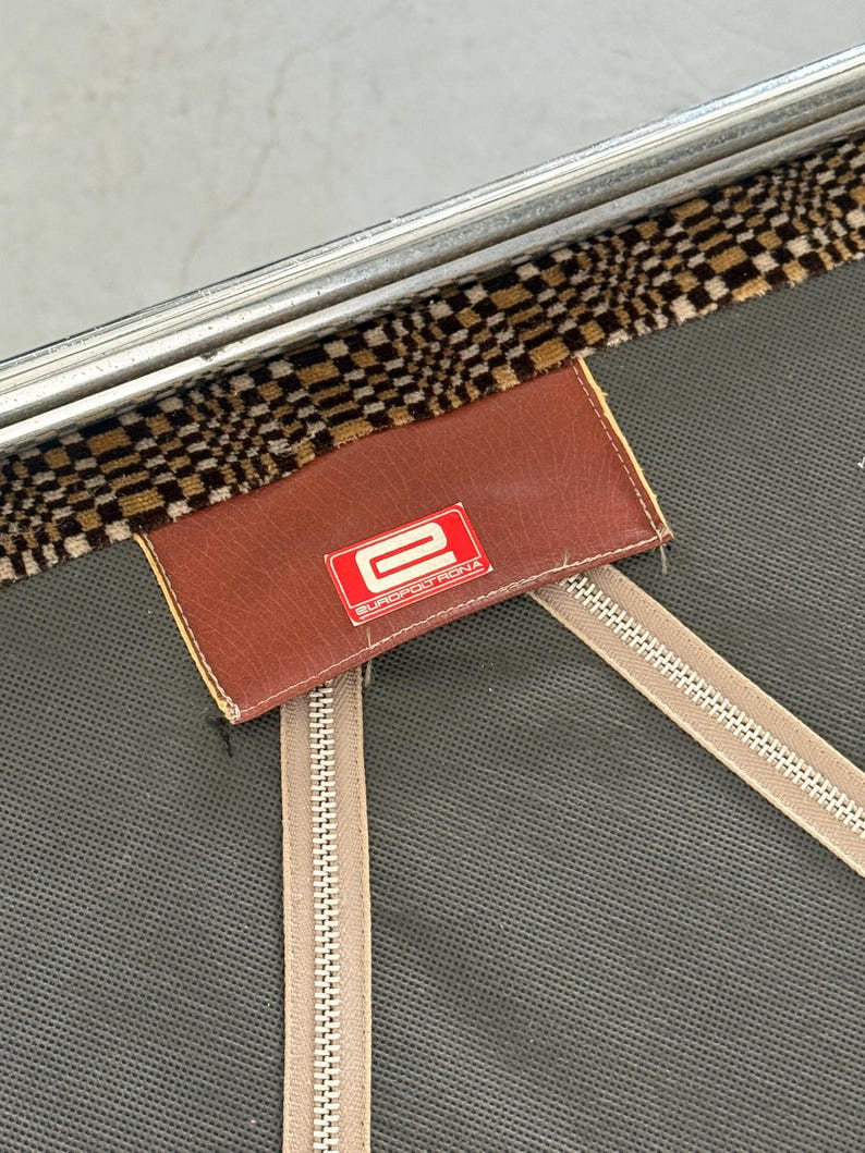 May include: Close-up of a vintage suitcase detail. The image shows a brown leather tag with the red "E" logo of EUROPOLTRONA. The tag is stitched onto a gray fabric with two beige zippers. A checkered brown and tan fabric trim is visible.