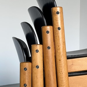 May include: Close-up of a wooden chair with a black backrest. The chair features light brown, cylindrical legs with visible wood grain and black screws. The backrest is composed of several curved, black wooden slats.