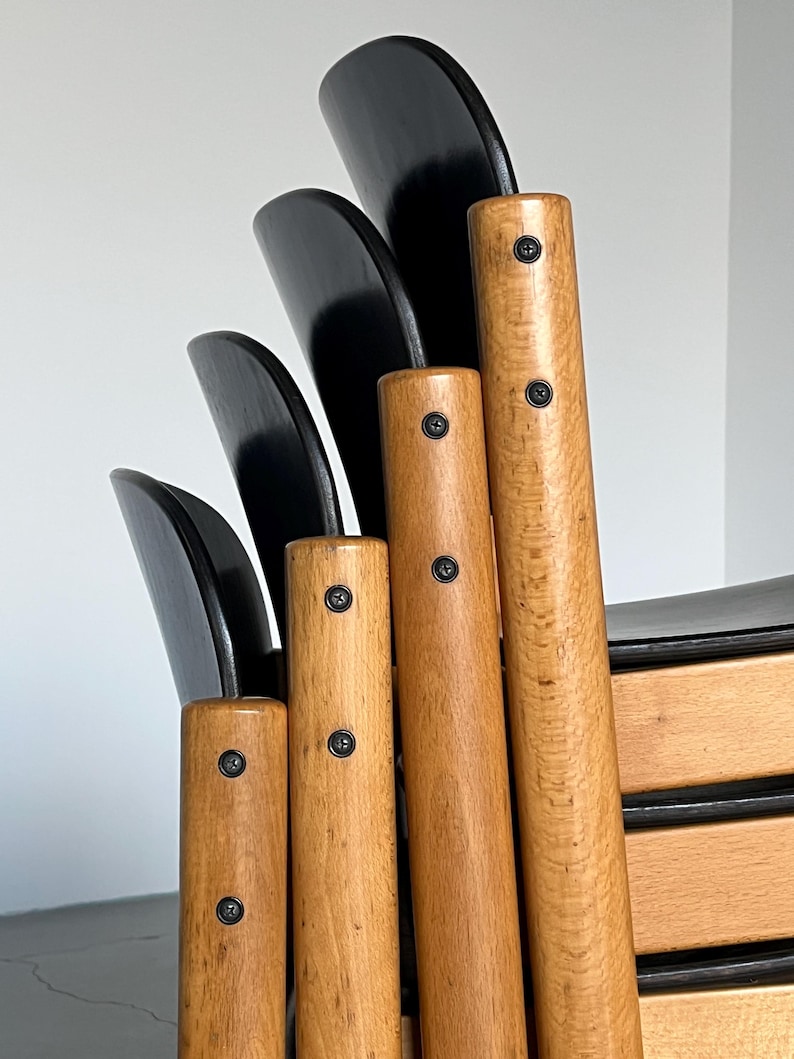 May include: Close-up of a wooden chair with a black backrest. The chair features light brown, cylindrical legs with visible wood grain and black screws. The backrest is composed of several curved, black wooden slats.
