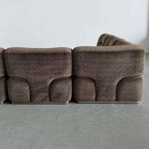 May include: A modular brown sofa with a geometric pattern. The sofa has a low profile with rounded edges and a metal base. The fabric has a textured appearance, and the design is modern and stylish.
