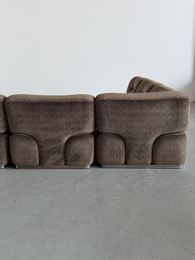 May include: A modular brown sofa with a geometric pattern. The sofa has a low profile with rounded edges and a metal base. The fabric has a textured appearance, and the design is modern and stylish.