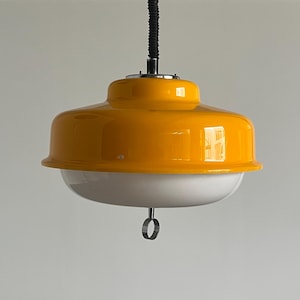 May include: A retro-style pendant light with a bright yellow, rounded shade and a white, bowl-shaped diffuser. The light hangs from a black, coiled cord and has a chrome-colored fixture. A small, circular ring hangs from the bottom.