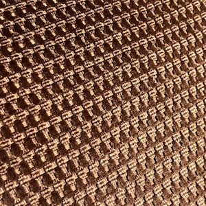 May include: Close-up of a textured fabric with a repeating pattern. The fabric is a warm brown color with a raised, woven design. The pattern creates a grid-like appearance, with each square having a slightly lighter shade.