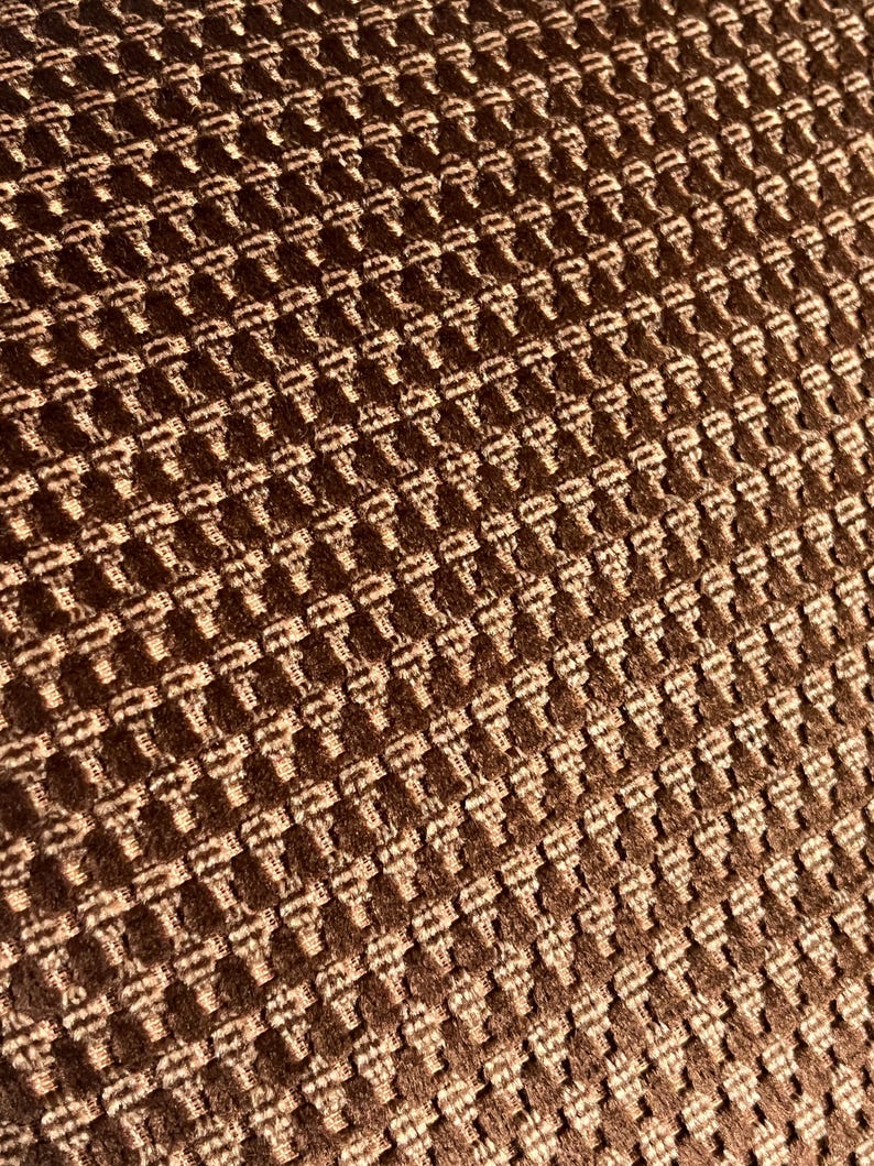 May include: Close-up of a textured fabric with a repeating pattern. The fabric is a warm brown color with a raised, woven design. The pattern creates a grid-like appearance, with each square having a slightly lighter shade.