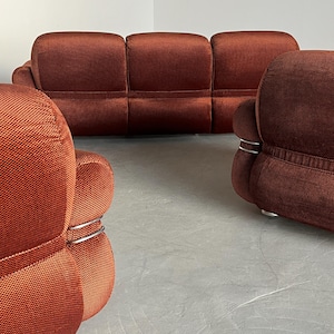 May include: A burnt orange three-piece modular sofa set. The sofa features a textured fabric upholstery and sleek, silver metal accents. The set includes a three-seater sofa and two armchairs. The furniture is arranged on a light gray floor.
