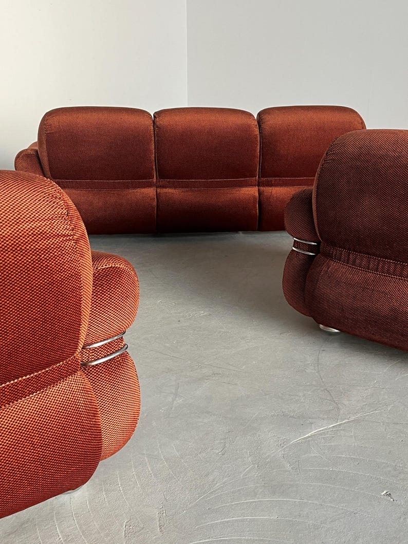 May include: A burnt orange three-piece modular sofa set. The sofa features a textured fabric upholstery and sleek, silver metal accents. The set includes a three-seater sofa and two armchairs. The furniture is arranged on a light gray floor.