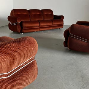 May include: A three-piece burnt orange velvet living room set, including a sofa and two armchairs. The furniture features a retro design with curved lines and metallic accents. The upholstery is a textured, deep orange velvet. The sofa has three seats and the armchairs have a single seat each. The pieces are arranged in a spacious room with a light-colored floor.