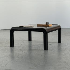 Könnte beinhalten: Ein niedriger, quadratischer Couchtisch mit glänzend schwarzer Oberfläche und abgerundeten, röhrenförmigen Kanten. Der Tisch hat vier stabile, zylindrische Beine und eine glatte, reflektierende Platte. Eine kleine Holzbox und Papiere liegen darauf.