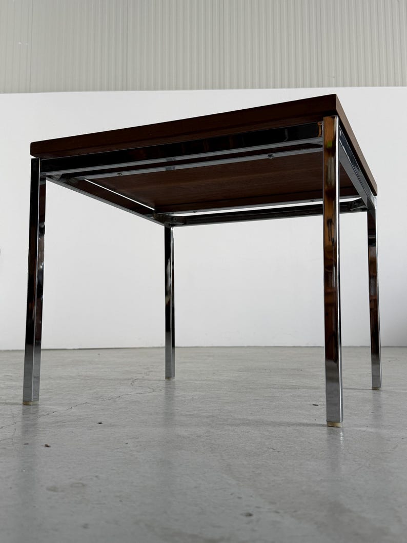 Op de afbeelding: Een bijzettafel in mid-century modern stijl met een donkerbruin houten blad en een verchroomd stalen frame. De tafel is vierkant, ongeveer 46 cm hoog en 61 cm breed.