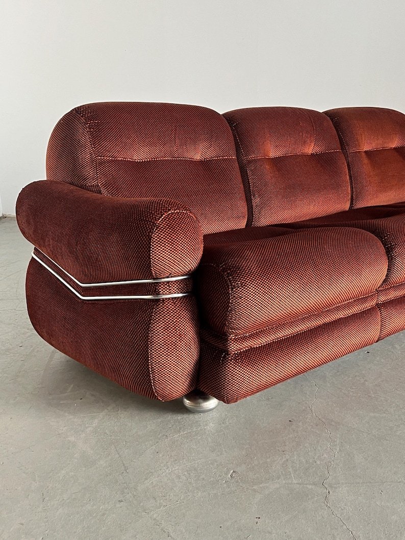 May include: A three-seater mid-century modern sofa upholstered in rust-colored corduroy. The sofa features a unique design with rounded armrests and sleek, metallic accents along the sides. The corduroy fabric has a subtle textured pattern, and the sofa sits on small, metallic legs. This vintage sofa is a statement piece that would add a touch of retro style to any living room.