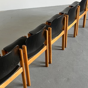 May include: A row of mid-century modern chairs. Each chair has a black seat and backrest, supported by a light-colored wooden frame. The chairs are arranged in a line, showcasing their design and construction. The chairs are likely made of wood and other materials.