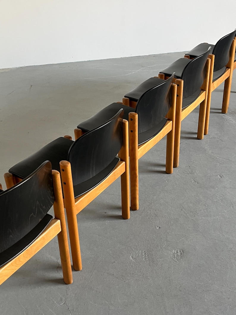May include: A row of mid-century modern chairs. Each chair has a black seat and backrest, supported by a light-colored wooden frame. The chairs are arranged in a line, showcasing their design and construction. The chairs are likely made of wood and other materials.
