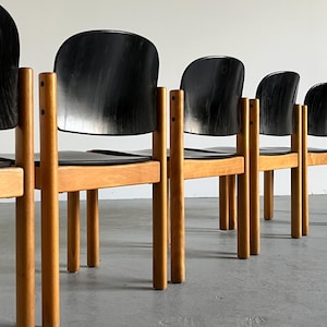 May include: A row of mid-century modern chairs. Each chair has a black, curved back and seat, supported by a light-colored wooden frame and legs. The chairs are arranged in a line against a neutral background.